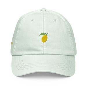 May include: A light green baseball cap with an embroidered lemon design. The lemon is yellow with a green leaf.
