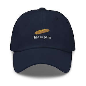 May include: A navy blue baseball cap with an embroidered image of a loaf of bread and the text "life is pain".