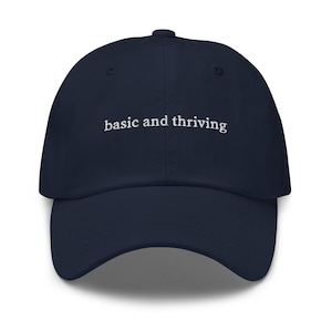 Puede incluir: Una gorra de béisbol azul marino con el texto "basic and thriving" bordado en blanco en la parte delantera.