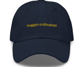 May include: A navy blue baseball cap featuring the embroidered words "nugget enthusiast" in gold. The cap has a curved peak and a classic six-panel design. A casual accessory.