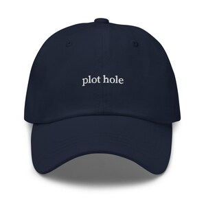 May include: A navy blue baseball cap featuring the words "plot hole" embroidered in white thread across the front center. The cap has a structured crown, a curved brim, and a button at the top, presenting a clean and simple design for headwear.