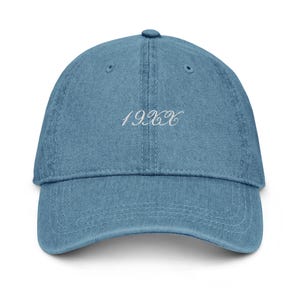May include: A light blue denim baseball cap with a curved peak. The cap has the embroidered text "19XX" in white script on the front. It features a six-panel design and an adjustable strap at the back.