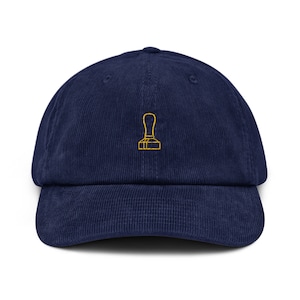 May include: A navy blue corduroy baseball cap with a yellow embroidered image of a coffee tamper.