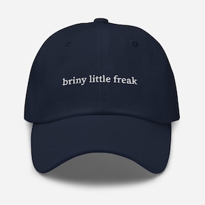 Op de afbeelding: Marineblauwe baseballcap met de witte tekst "briny little freak" geborduurd op de voorkant. De cap heeft een gebogen klep en een knoop aan de bovenkant. Een casual accessoire.