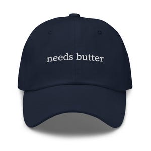 May include: A navy blue baseball cap with white embroidered text that reads "needs butter".