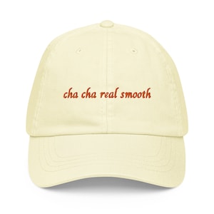 May include: A cream-coloured baseball cap featuring the embroidered words "cha cha real smooth" in reddish-orange thread. The cap has a curved peak and a classic six-panel design.
