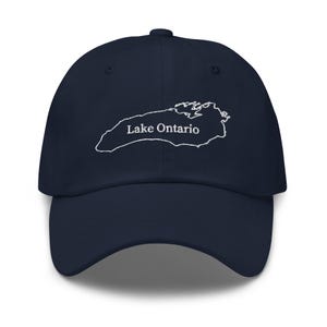 May include: A navy blue baseball cap featuring a white embroidered outline of Lake Ontario on the front panel. The text Lake Ontario is stitched in white lettering directly across the center of the lake map graphic on the crown of the hat.