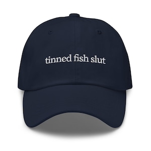 May include: A navy blue baseball cap with the embroidered text "tinned fish slut" in white. The cap has a curved brim and a button on top. This hat is a casual accessory.