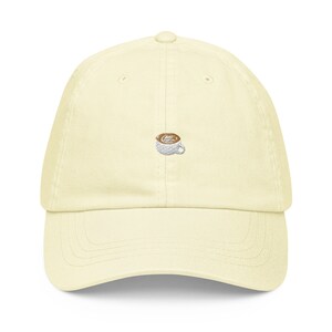 Puede incluir: Una gorra de béisbol de color amarillo claro con una imagen bordada de una taza de café con un líquido marrón.
