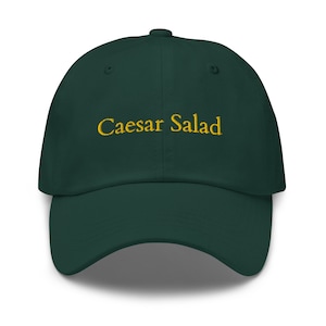 Puede incluir: Una gorra de béisbol verde oscuro con el texto "Caesar Salad" bordado en amarillo.