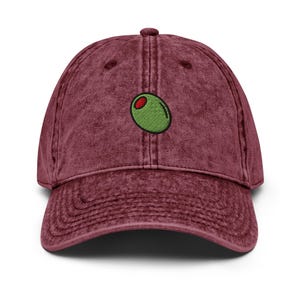 May include: A maroon baseball cap with a pre-washed, vintage look. The cap features an embroidered green olive with a red center. The cap has a curved brim and an adjustable strap for a comfortable fit. This hat is a fun accessory for casual wear.