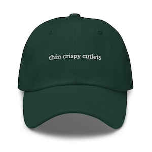 May include: A dark green baseball cap with the text "thin crispy cutlets" embroidered in white on the front.
