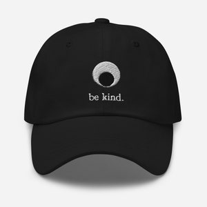 Może przedstawiać: Czarna czapka z daszkiem z białym kółkiem i haftowanym napisem "be kind."