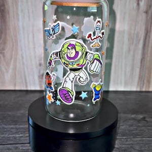 K&ouml;nnte beinhalten: Ein klares Trinkglas mit Holzdeckel, verziert mit Aufklebern von Zeichentrickfiguren. Das Glas zeigt Buzz Lightyear und andere Charaktere aus dem Toy Story-Franchise. Das Glas wird auf einem schwarzen St&auml;nder pr&auml;sentiert.