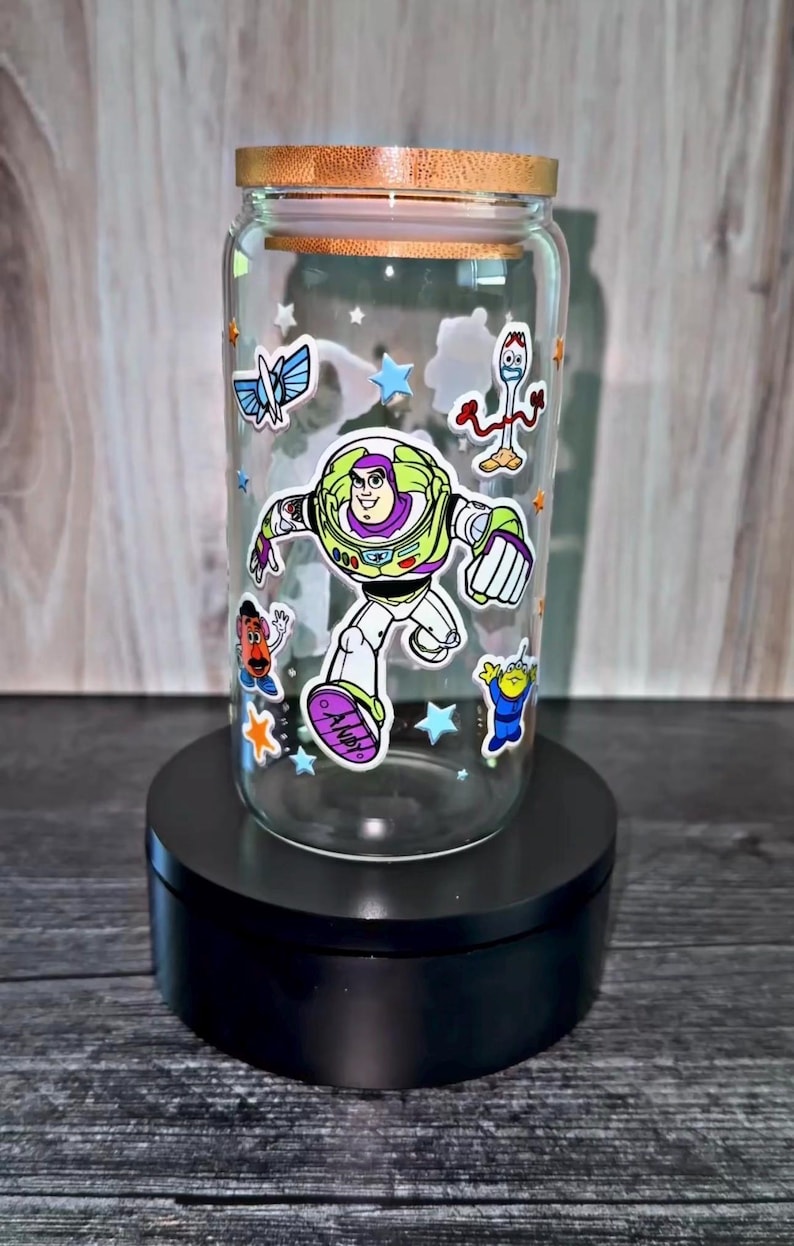 K&ouml;nnte beinhalten: Ein klares Trinkglas mit Holzdeckel, verziert mit Aufklebern von Zeichentrickfiguren. Das Glas zeigt Buzz Lightyear und andere Charaktere aus dem Toy Story-Franchise. Das Glas wird auf einem schwarzen St&auml;nder pr&auml;sentiert.