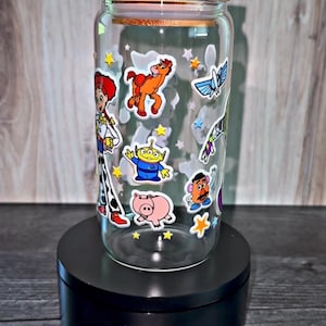 K&ouml;nnte beinhalten: Ein durchsichtiger Glasbecher mit Holzdeckel, verziert mit bunten Aufklebern von Figuren aus dem Animationsfilm Toy Story. Der Becher steht auf einem schwarzen St&auml;nder und zeigt das verspielte Design.