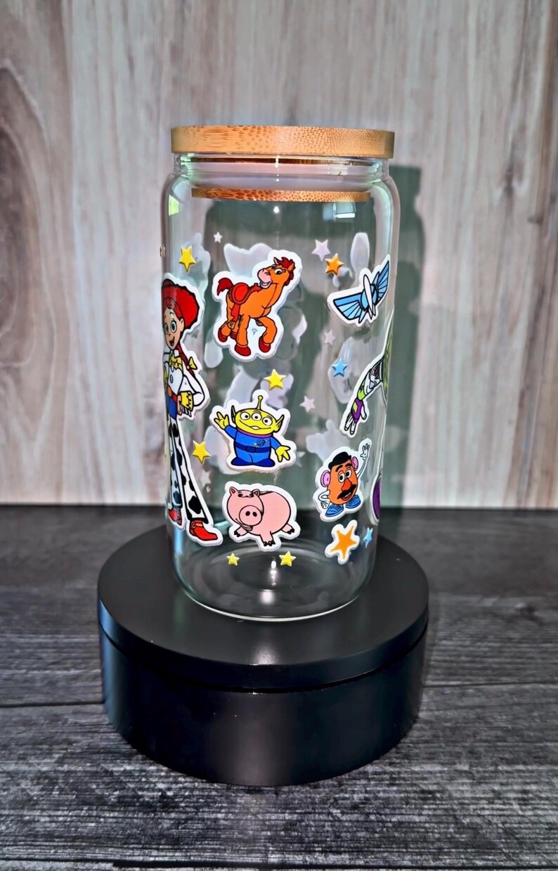 K&ouml;nnte beinhalten: Ein durchsichtiger Glasbecher mit Holzdeckel, verziert mit bunten Aufklebern von Figuren aus dem Animationsfilm Toy Story. Der Becher steht auf einem schwarzen St&auml;nder und zeigt das verspielte Design.