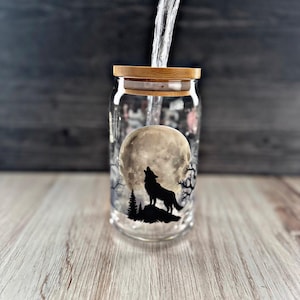 May include: A glass jar with a wooden lid, featuring a black silhouette of a wolf howling at the moon. The jar is filled with water, and the moon is depicted as a full moon.