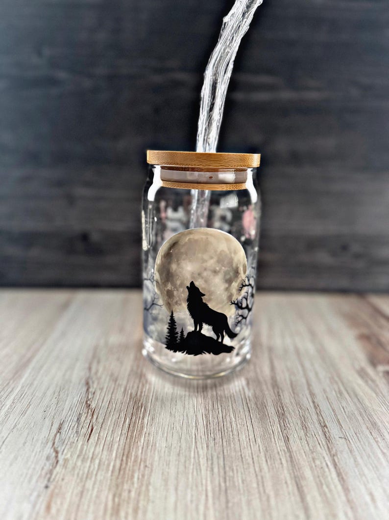 May include: A glass jar with a wooden lid, featuring a black silhouette of a wolf howling at the moon. The jar is filled with water, and the moon is depicted as a full moon.