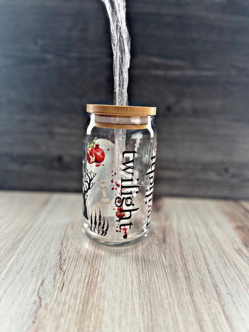 May include: A clear glass jar with a wooden lid and a black and white design featuring the word "twilight" and a red pomegranate. The jar is filled with a clear liquid and a clear plastic straw.