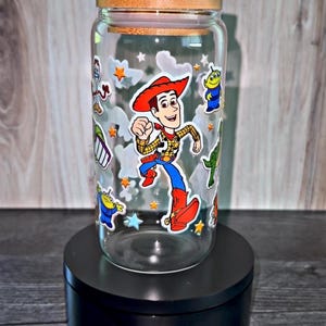 K&ouml;nnte beinhalten: Ein klares Glas mit einem Holzdeckel, verziert mit Aufklebern von Zeichentrickfiguren. Das Glas zeigt ein Woody-Charakterdesign sowie andere Charaktere und Sternaufkleber. Das Glas wird auf einem schwarzen St&auml;nder ausgestellt.