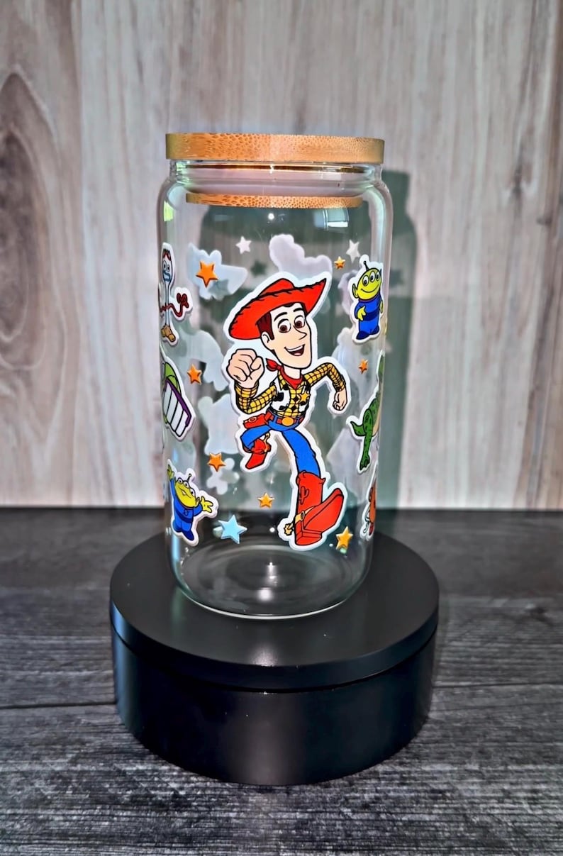 K&ouml;nnte beinhalten: Ein klares Glas mit einem Holzdeckel, verziert mit Aufklebern von Zeichentrickfiguren. Das Glas zeigt ein Woody-Charakterdesign sowie andere Charaktere und Sternaufkleber. Das Glas wird auf einem schwarzen St&auml;nder ausgestellt.