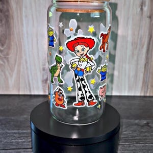 K&ouml;nnte beinhalten: Ein klares Glas mit einem Holzdeckel, verziert mit Zeichentrickfiguren aus dem Film Toy Story. Das Glas zeigt Bilder von Jessie, den Aliens und anderen Charakteren. Das Glas steht auf einem schwarzen St&auml;nder.