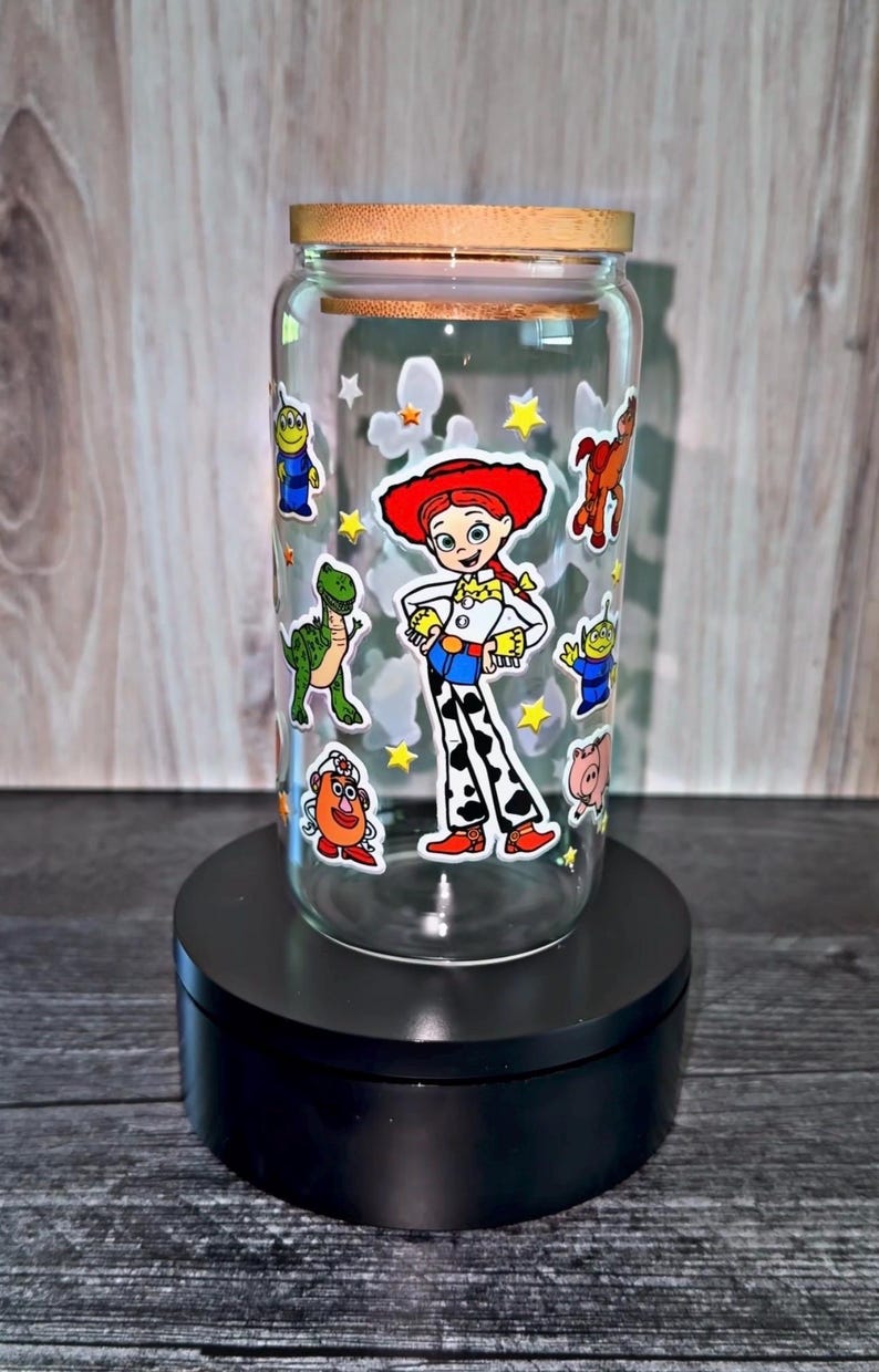 K&ouml;nnte beinhalten: Ein klares Glas mit einem Holzdeckel, verziert mit Zeichentrickfiguren aus dem Film Toy Story. Das Glas zeigt Bilder von Jessie, den Aliens und anderen Charakteren. Das Glas steht auf einem schwarzen St&auml;nder.
