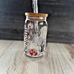 May include: A clear glass jar with a gold lid and a black and white design featuring a tree, a paw print, a chess piece, and the word "Twilight". The jar contains a red pomegranate.