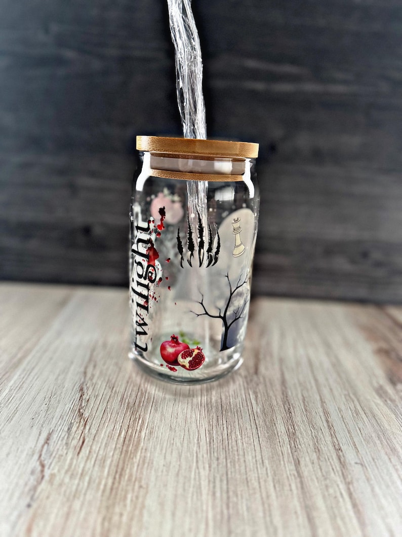 May include: A clear glass jar with a gold lid and a black and white design featuring a tree, a paw print, a chess piece, and the word "Twilight". The jar contains a red pomegranate.