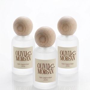 May include: Three frosted glass bottles with round wooden stoppers. Each bottle features a cream-colored label with the brown text Olivia & Morgan and the date 18th September 2026 printed in a serif font. The bottles have a cylindrical shape.