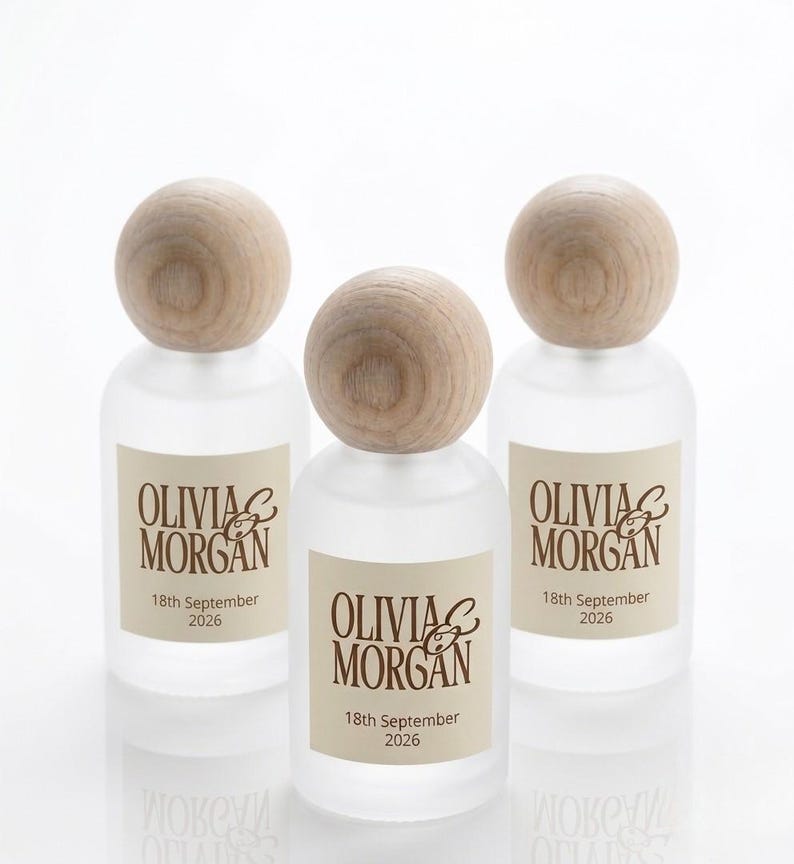 May include: Three frosted glass bottles with round wooden stoppers. Each bottle features a cream-colored label with the brown text Olivia & Morgan and the date 18th September 2026 printed in a serif font. The bottles have a cylindrical shape.