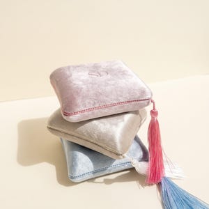 Puede incluir: Una pila de tres cajas cuadradas de terciopelo para anillos en tonos rosa claro, beige y azul claro. La caja superior presenta las letras SD en relieve. Cada caja está rematada con un cordón trenzado a juego y una larga borla de seda.