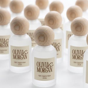 May include: A collection of small, frosted glass bottles with round, light-colored wooden stoppers. Each bottle features a rectangular label with the text "OLIVIA & MORGAN" printed in brown, followed by the date "18th September 2026" below the names.