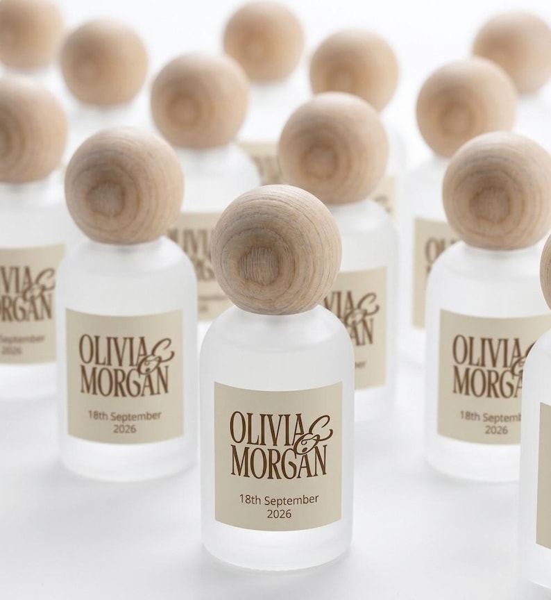 May include: A collection of small, frosted glass bottles with round, light-colored wooden stoppers. Each bottle features a rectangular label with the text "OLIVIA & MORGAN" printed in brown, followed by the date "18th September 2026" below the names.