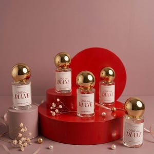 Op de afbeelding: Een verzameling parfumflesjes met gouden bolvormige doppen en witte etiketten met de tekst "With love DIANE". De flesjes staan op rode en roze sokkels met decoratieve elementen, waaronder parels en droogbloemen.