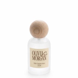 May include: A frosted glass bottle with a round, light-colored wooden stopper. A rectangular cream-colored label on the front features the brown text OLIVIA & MORGAN and the date 18th September 2026 printed in a clear, dark brown font.