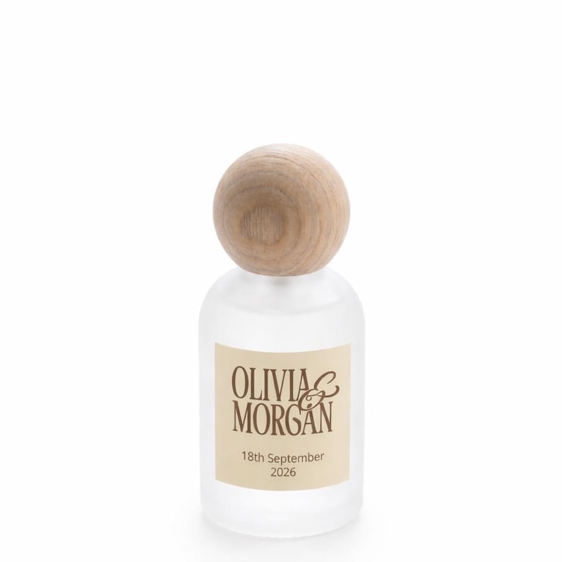 May include: A frosted glass bottle with a round, light-colored wooden stopper. A rectangular cream-colored label on the front features the brown text OLIVIA & MORGAN and the date 18th September 2026 printed in a clear, dark brown font.