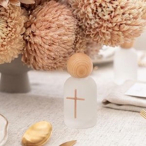May include: A small, cylindrical, frosted glass bottle featuring a light brown wooden sphere as a stopper. A simple, thin, light brown cross is printed on the front of the bottle. The bottle is positioned upright on a textured, light-colored surface.