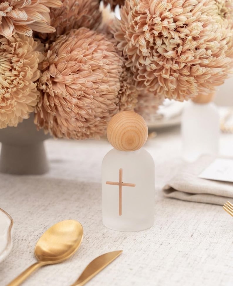 May include: A small, cylindrical, frosted glass bottle featuring a light brown wooden sphere as a stopper. A simple, thin, light brown cross is printed on the front of the bottle. The bottle is positioned upright on a textured, light-colored surface.