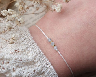 May include: Delicate white string bracelet featuring two small, light blue beads and two gold-colored beads. The bracelet is simple and elegant, perfect for everyday wear. The bracelet is shown on a wrist.
