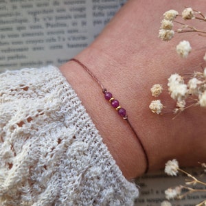 Könnte beinhalten: Ein schlichtes Armband mit einem braunen Band und drei kleinen, dunkelrosa Perlen. Das Armband wird am Handgelenk getragen.