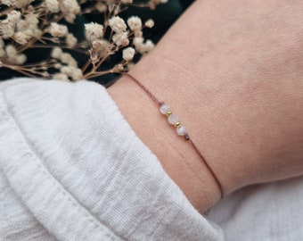 May include: A simple bracelet with three small white stones and gold accents on a brown cord.