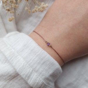 Könnte beinhalten: Ein zartes Armband mit einem kleinen violetten Edelstein und goldenen Akzenten auf einem braunen Band.