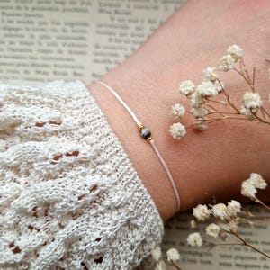 Könnte beinhalten: Ein weißes Armband aus Schnur mit einer kleinen grauen und weißen Perle. Das Armband befindet sich an einem Handgelenk und der Hintergrund ist ein Buch mit weißen Blumen.