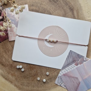 May include: A delicate bracelet with a crescent moon charm and white beads on a brown string. The bracelet is displayed on a white card with a pink circle and a crescent moon design.