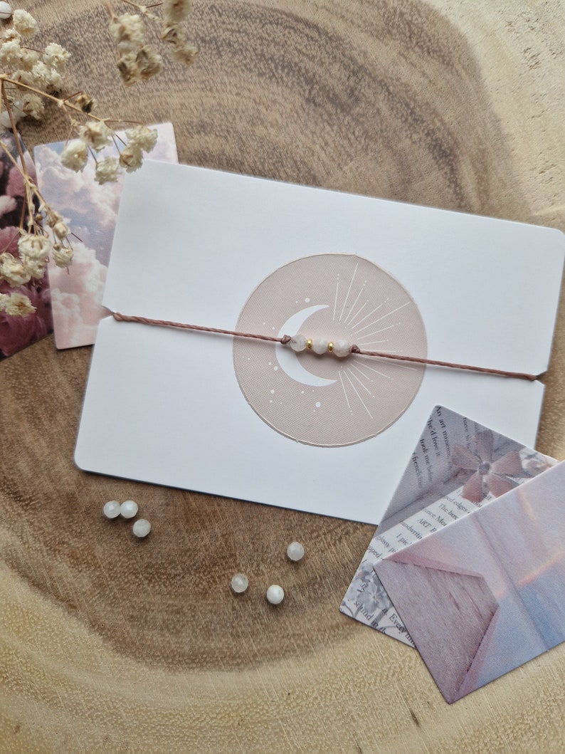 May include: A delicate bracelet with a crescent moon charm and white beads on a brown string. The bracelet is displayed on a white card with a pink circle and a crescent moon design.