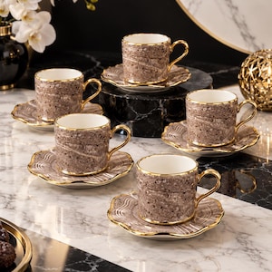 Puede incluir: Un juego de cuatro tazas de café y platillos con un patrón de mármol moteado en marrón y blanco, con bordes y asas en tono dorado. Cada taza tiene una forma cilíndrica y cada platillo presenta un borde festoneado con un diseño a juego.