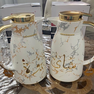 May include: Two white thermal carafes with gold accents and marble-like patterns. The carafes have gold lettering on them, one says "Shay" and the other says "Always".