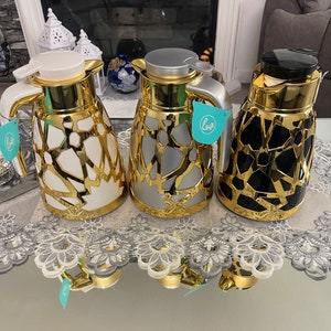 May include: Three gold-coloured thermal carafes with different coloured lids and bodies. The carafes have a geometric pattern and are on a white lace tablecloth. The text "عش" is on the carafes.